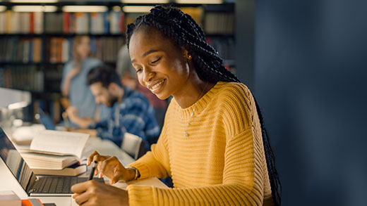 Smiling young woman with braided hair studying on her laptop in a lively library setting, surrounded by fellow students immersed in their books, exemplifying a focused yet collaborative academic atmosphere.